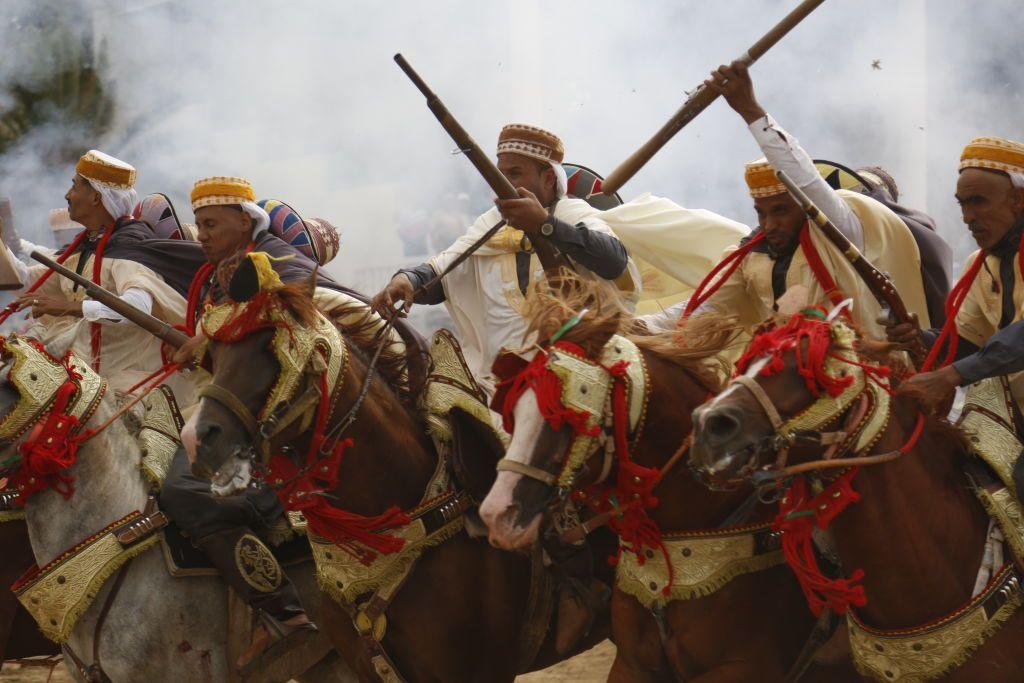 Men holding old-fashioned rifles ride at speed in formation. They are wearing matching robes, and the the horses they are riding have matching elaborate bridles.