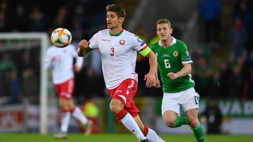 A Belarusian footballer, wearing a white shirt, red shorts and white socks chases a football. He is chased by a Northern Ireland player in a green shirt, white shorts and green socks