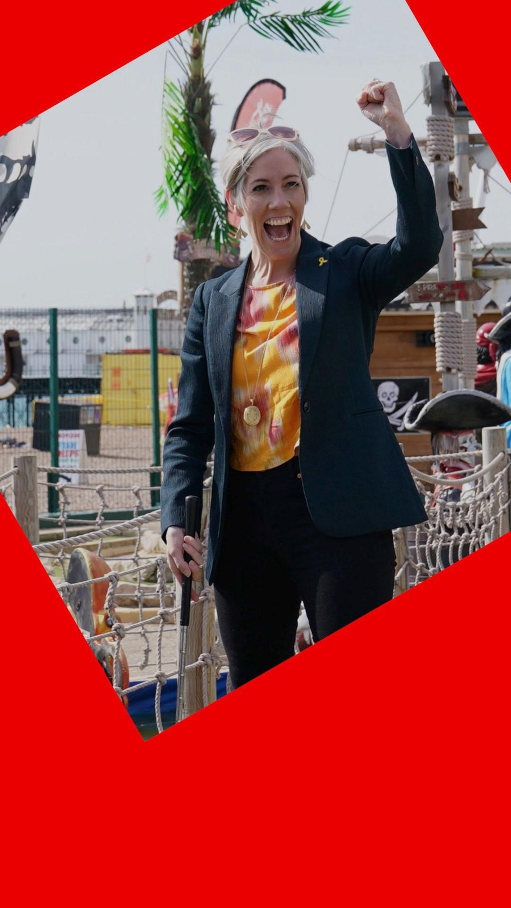 A woman, Liberal Democrat deputy leader Daisy Cooper, smiles while playing crazy golf - she wears a dark suit and her left arm is raised