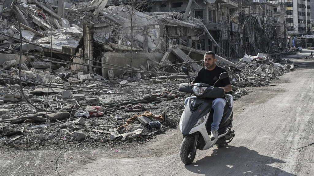 A man in Lebanon drives his moped along a gravel path, next to a building that has been destroyed by an Israeli air strike