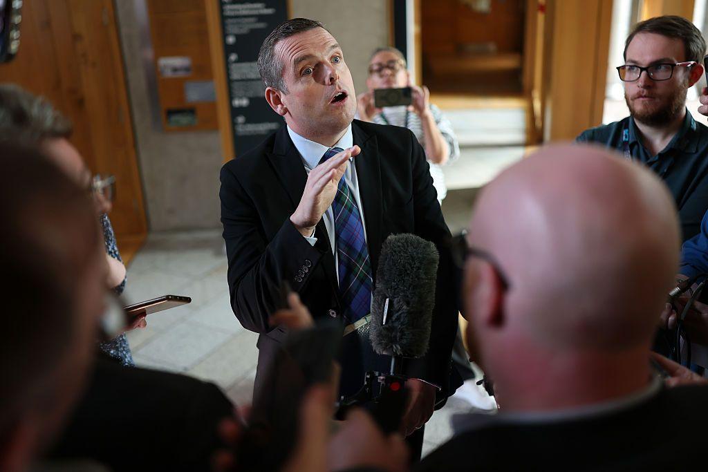 Douglas Ross in blue and green tartan tie, a dark suit and a light shirt is speaking to journalists in the lobby of the Holyrood building. He is caught mid-comment, gesticulating with his right hand and speaking to someone off camera. A crowd of journalists surrounds him, holding up mics and cameras.