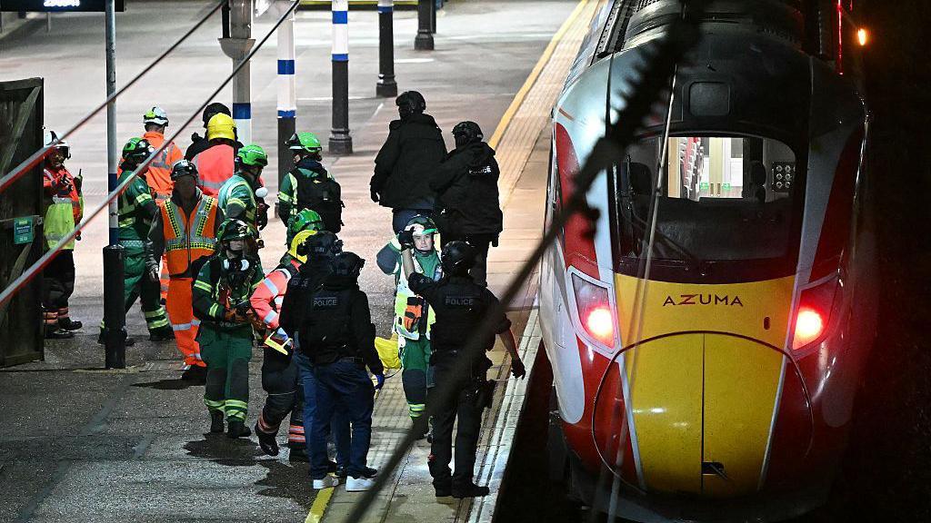 Black-clad police officers and emergency crews wearing green-and-yellow coveralls on the platform of Huntingdon train station, where a train has stopped