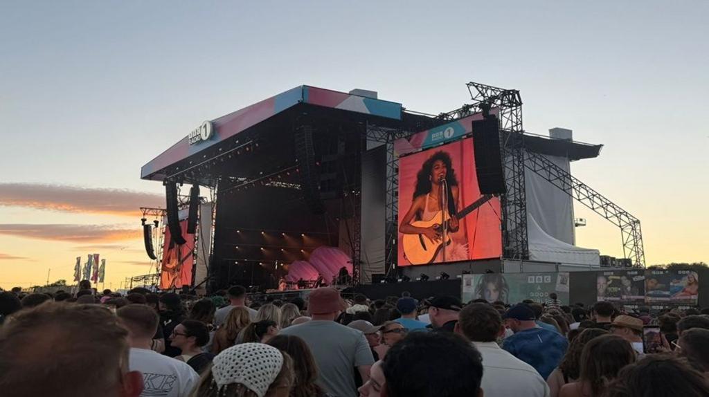A wide shot of the stage at Big Weekend with Olivia Dean on stage and projected on two large screens at either side of the stage. In the foreground is the crowd with many of them holding up phones
