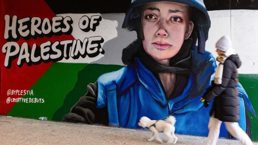 A member of the public walks a dog past pro-Palestinian street art in tribute to Plestia Alaqad in an underpass. On the mural, there are the words 'Heroes of Palestine'. Plestia is wearing a blue press-jacket in the mural.
