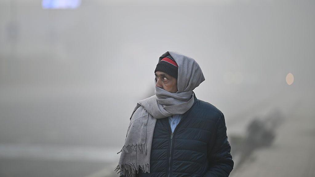 A man wearing a black jacket and a red-and-black woollen headgear uses a grey muffler to cover his ears, mouth and nostrils as he walks through a thick haze - which has almost blurred everything behind him - in the streets of India's Delhi