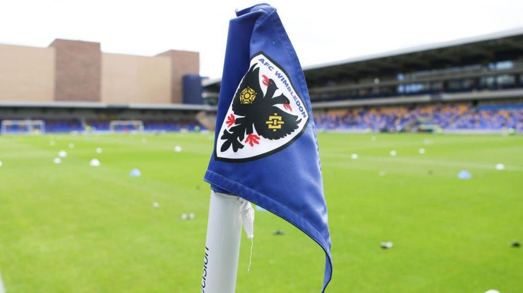 A corner flag at the Cherry Red Records Stadium with AFC Wimbledon branding on it, with the pitch and stands in the background