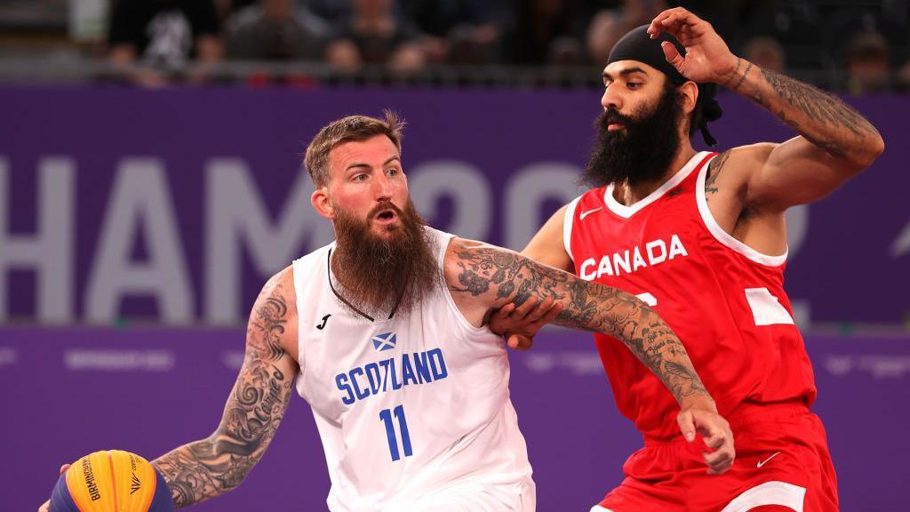A basketball player wearing a white Scotland jersey holds off another player in a red Canada jersey while on the court.