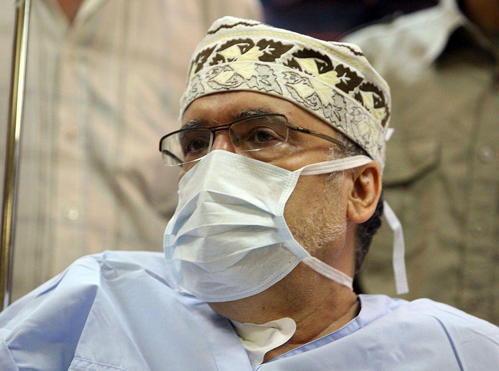 Abdelbaset Ali Mohamed al-Megrahi in hospital overalls wearing a mask and glasses and a small, patterned hat. We can see he has a beard and two men in light shirts are standing behind him.