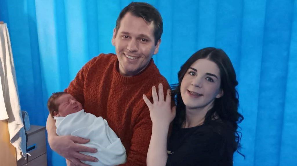 A man in a red pullover holds a dark haired baby while his partner, a dark haired woman in a black top, holds on to his shoulder and smiles at the camera.