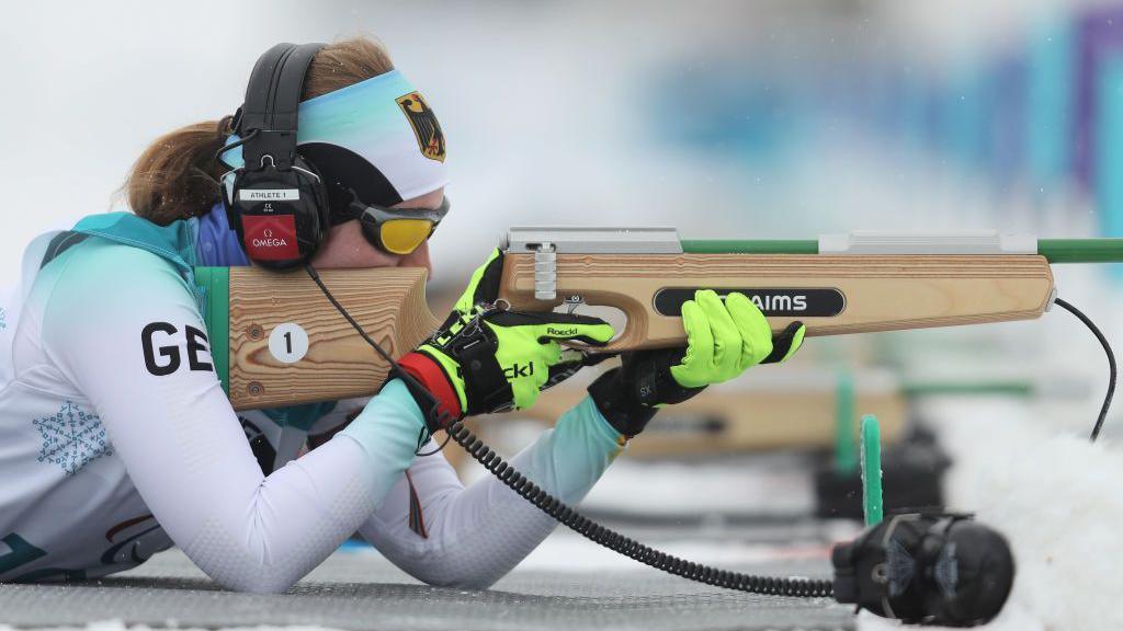 Germany's Clara Klug wears headphones to hear acoustic signals to find the target while shooting her rifle while lying prone on the floor in the biathlon