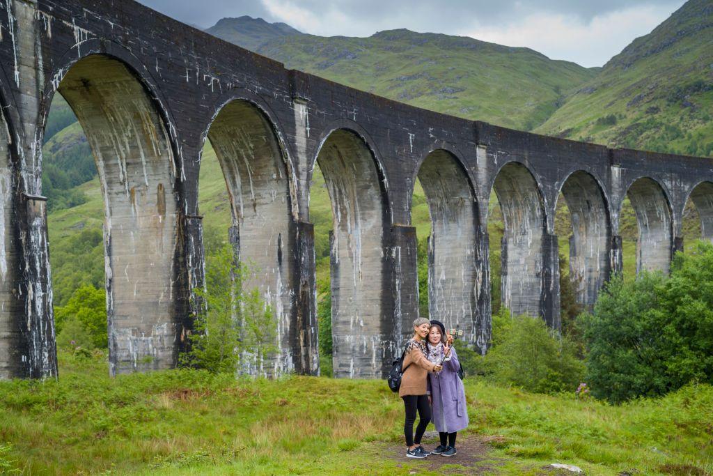 Two tourists stand in front of Glenfinnan Viaduct, a dramatic railway arch in the West Highlands in Scotland. There are taking a selfie with a selfie stick