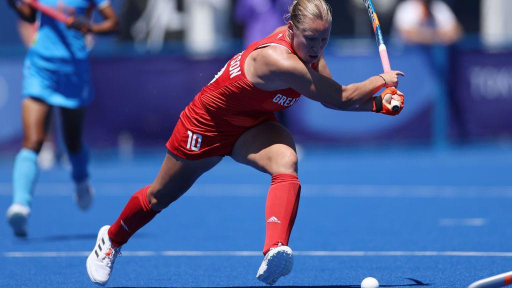 Hockey player Sarah Robertson, with blond hair tied back and in red kit and white trainers on a blue hockey pitch, prepares to make a reverse strike, while playing for Great Britain