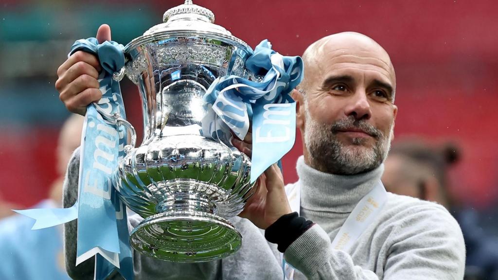 Pep Guardiola, wearing a grey polo neck jumper, holds the FA Cup aloft
