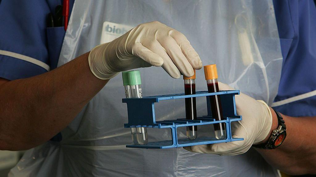 A Biobank employee wearing a blue nurse's uniform, white apron and white disposable gloves, handles vials of blood in a lab.