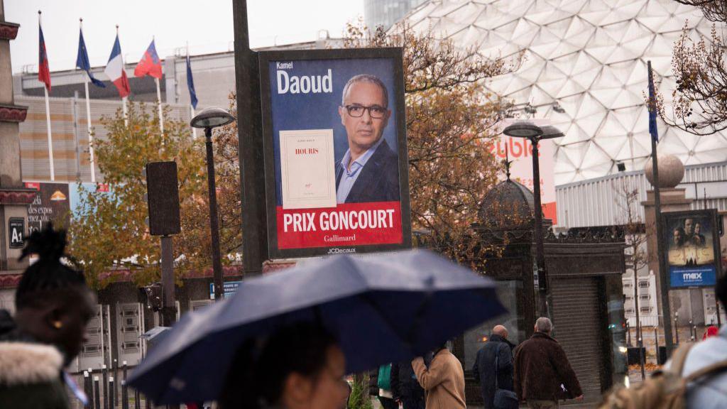 Parisians walk in the rain past a poster of Algerian writer Kamel Daoud, alongside his prize-winning book Houris