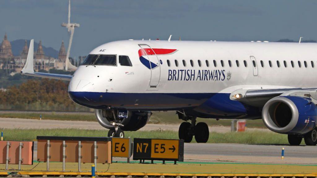 A British Airways plane sits on the runway in Barcelona, with signs on the ground and buildings in the background