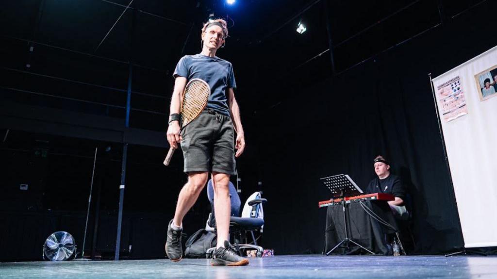 A man wearing shorts and a t-shirt holding a squash racket, on stage in a theatre.