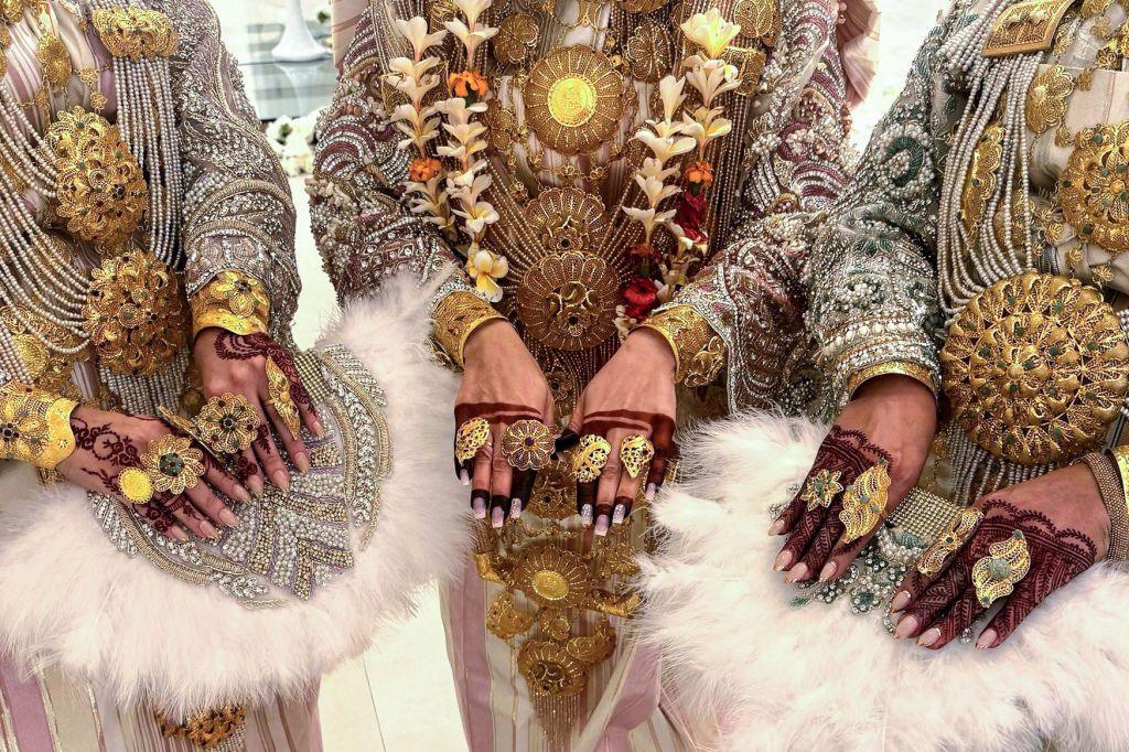 Women wear ornate golden necklaces and rings. On their fingers they have brown-red henna patterns.