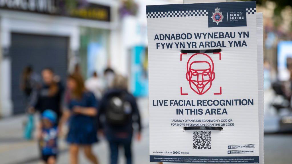 A close up of a poster in a city centre with people walking in the background. The poster says 'Live facial recognition in this area' with a barcode and an outline of facial structure. 