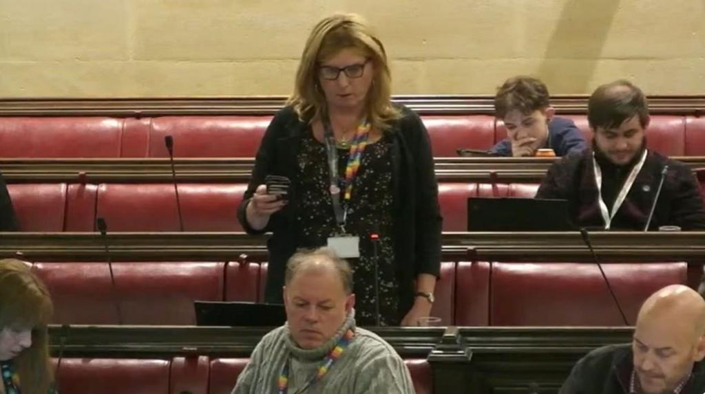 Kaz Self standing at a council meeting. She has blonde hair and is wearing a black dress and a black cardigan. Several other councillors are sitting on the benches.