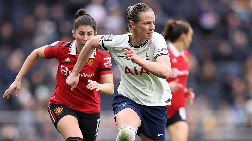 Two footballers running on a pitch. One woman is in a red and white Manchester United T-shirt and has brown hair in a bun. The other is wearing a white Tottenham Hotspur T-shirt with navy piping, and has light brown hair in a ponytail.