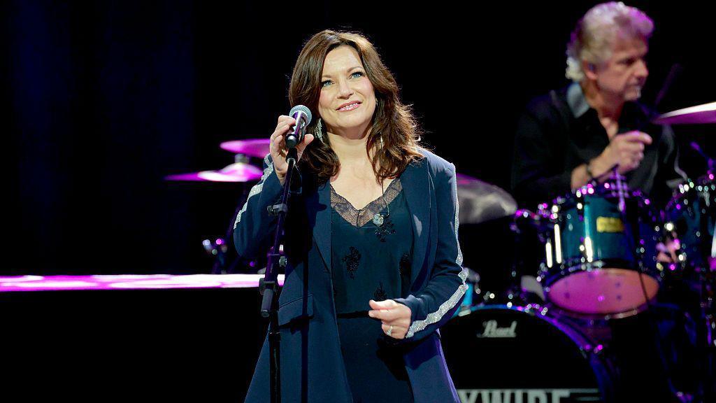 A woman with wavy brown hair in a black pant suit smiles in front of a microphone
