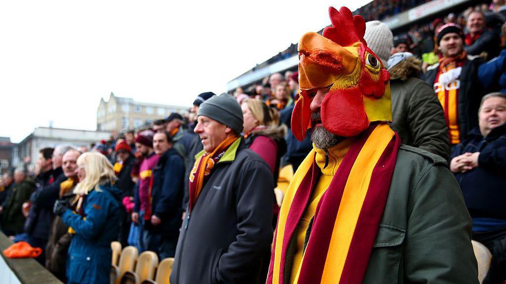 Bradford City fans stand up in the stands to watch a football match, many wear red and yellow club scarves and a man at the front wears a bantam mask 