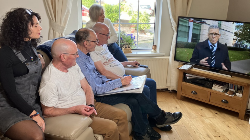 Show five people, including two women on either side of three men, sitting on a settee watching television