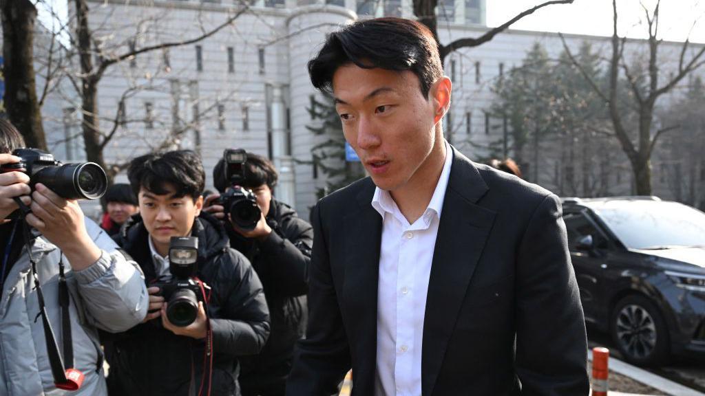 Hwang Ui-jo in a black suit, walking and being photographed by a group of men with cameras