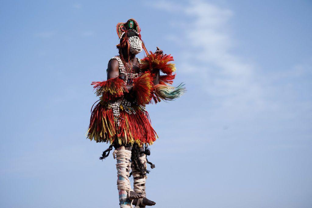 A man weaing an eye-catching outfit made of cowries and red-and-yellow tassels stands on stilts.