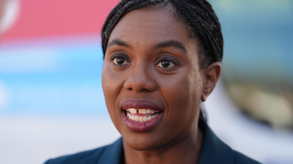 Head and shoulders shot of Kemi Badenoch looking at the camera and speaking. She is weraing a navy blue jacket, and there is a blurred light blue and red background.
