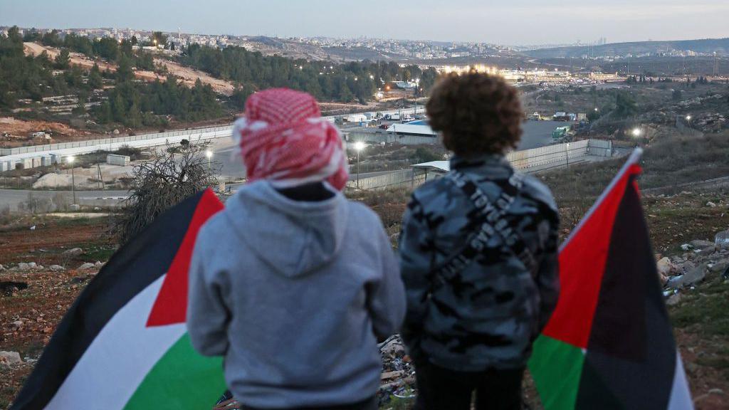 Two Palestinians with their backs to the camera hold black, white, green and red Palestinian flags while look down on Ofer prison, in the occupied West Bank, ahead of the release of dozens of Palestinian prisoners  in exchange for three Israeli hostages on the first day of the Gaza ceasefire deal (19 January 2025)