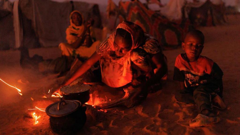 A woman in a refugee camp cooks at an open fire with the light from the flames illuminating her face. A child sits to her left. 