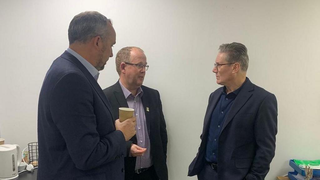 Kelvin Thomas, Tom Cliffe and Sir Keir Starmer inside a meeting room at the Sixfields stadium. The walls are grey and to the left is a kettle. All three men are wearing suits and engaged in conversation.