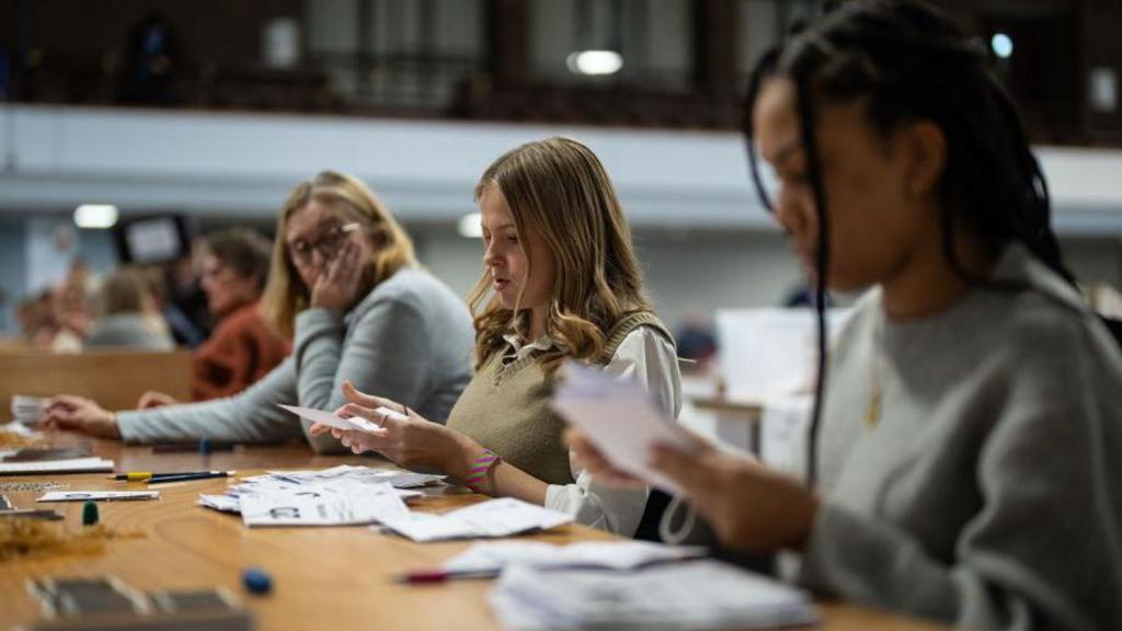 Three women counting ballot papers at a wooden desk. 