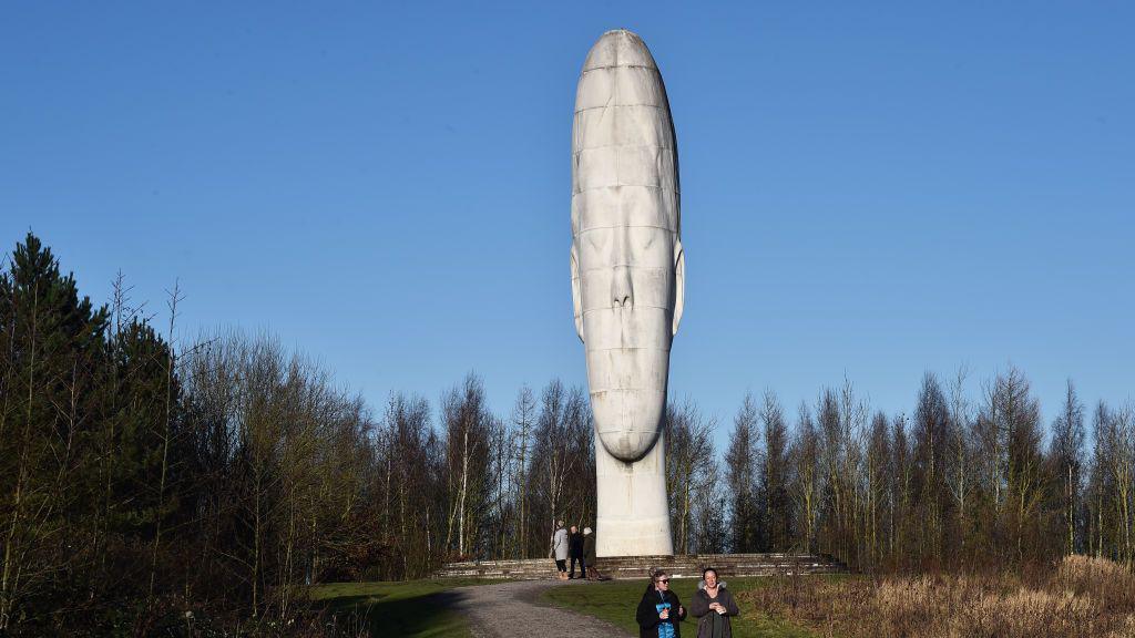 The Dream Sculpture, which is huge white stonework showing a young girl's head looming over the trees around it. Some people can be seen walking around the bottom of it on a green mound.
