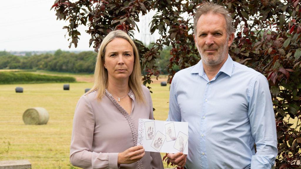 Sarah and Jack Hawkins hold up a piece of paper showing the hand and foot prints of their daughter Harriet. They are standing just off a field, a tree behind them