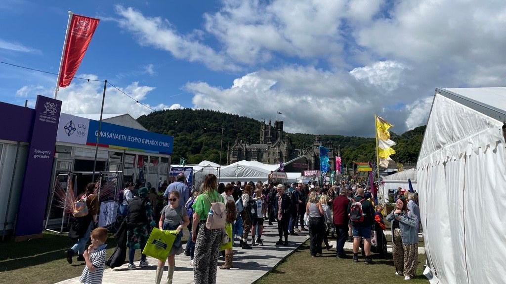 Crowds can be seen among two row of stalls in white tents on each side of a path at the Maes. Margam Castle can be seen in the background among green hills and blue skies. 