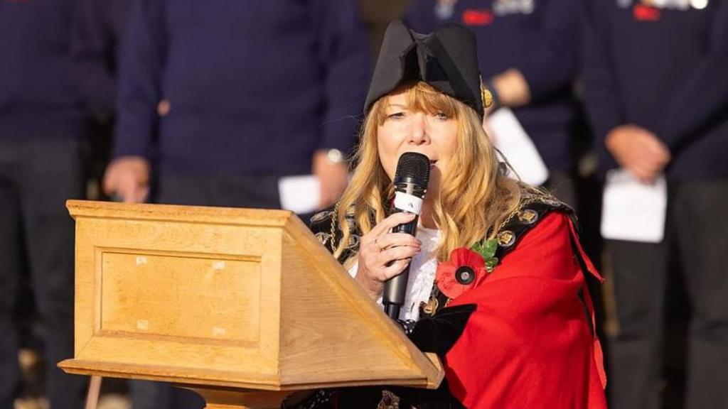 The mayor of Aldeburgh TJ Halworth-Culf - a woman with long ginger/blonde hair dressed in a red mayoral gown and wearing a black hat. She is holding a mic. 