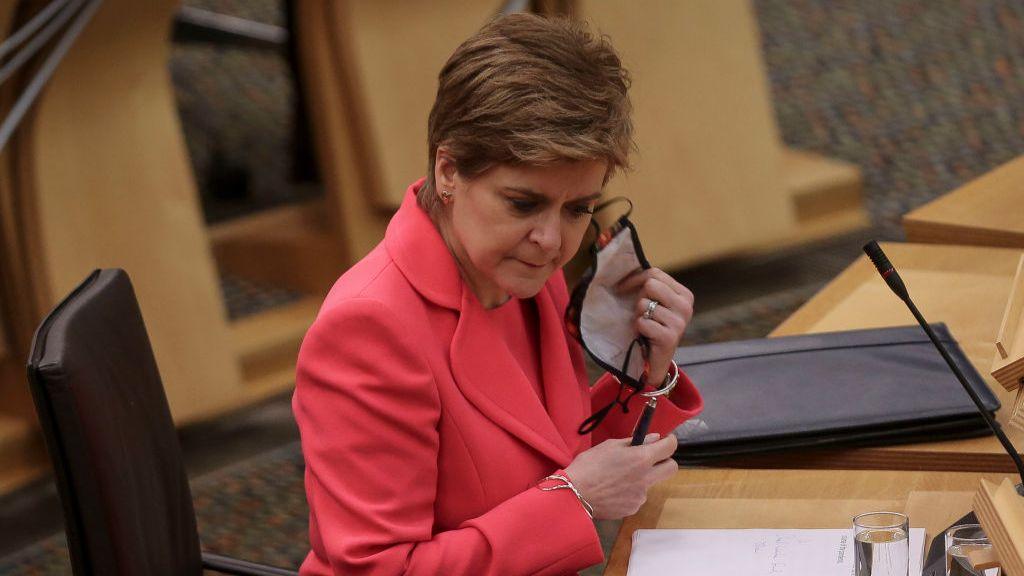 Nicola Sturgeon - a woman with short dark hair, wearing a pink jacket - pictured removing a face mask in the Scottish Parliament chamber during the pandemic