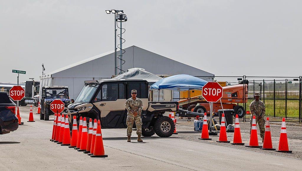 Men in military uniforms stand in between bright orange traffic cones and large signs saying 'STOP' with tents, vehicles and an airfield in the background - the view of Alligator Alcatraz from the outside.