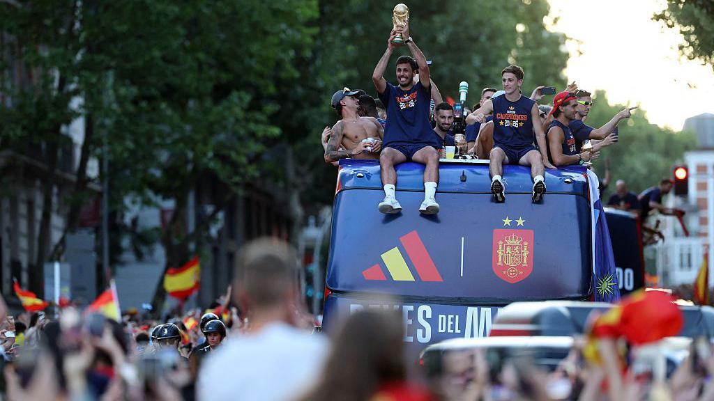 Rodri sits at the front on top of the bus holding the World Cup trophy above his head