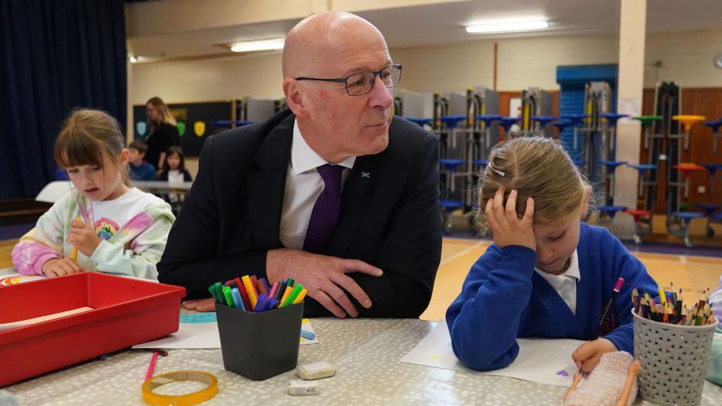 First Minister John Swinney visiting a school, sitting a table with two small girls who are doing some drawing. There are felt tip pens and coloured pencils in tubs on the table.