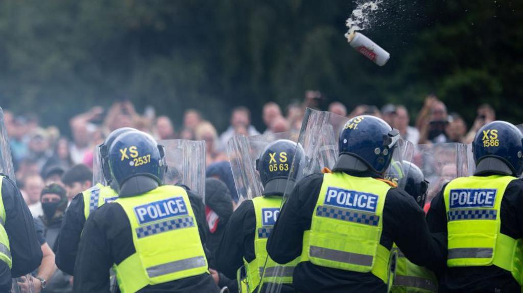 Point of view is standing behind police officers holding riot shields. A beer can is about to hit one of them. In the distance there are many blurred figures taking part in the "protest".