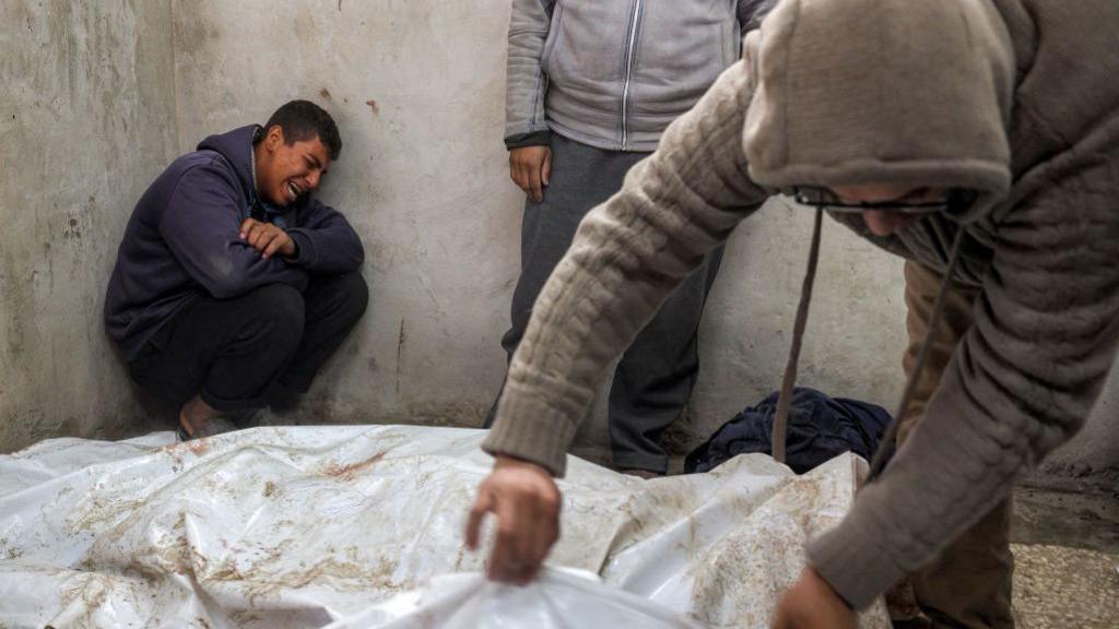 Boy cries in the background as a man looks at a shrouded corpse in gaza