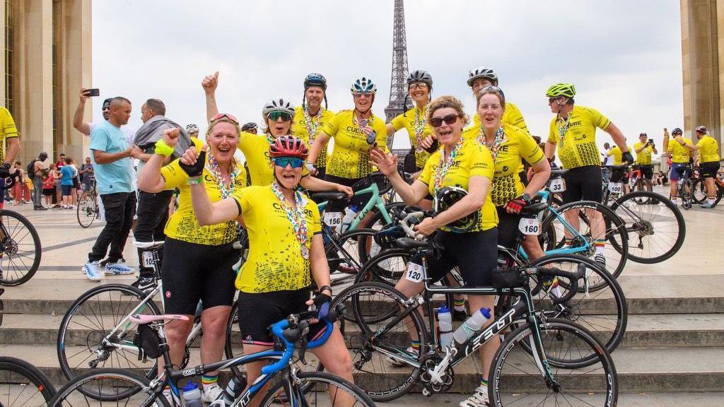 Angela Mills-Bannon and a group of cyclists in yellow jersey's stood in Paris with the Eiffel Tower in the distance.