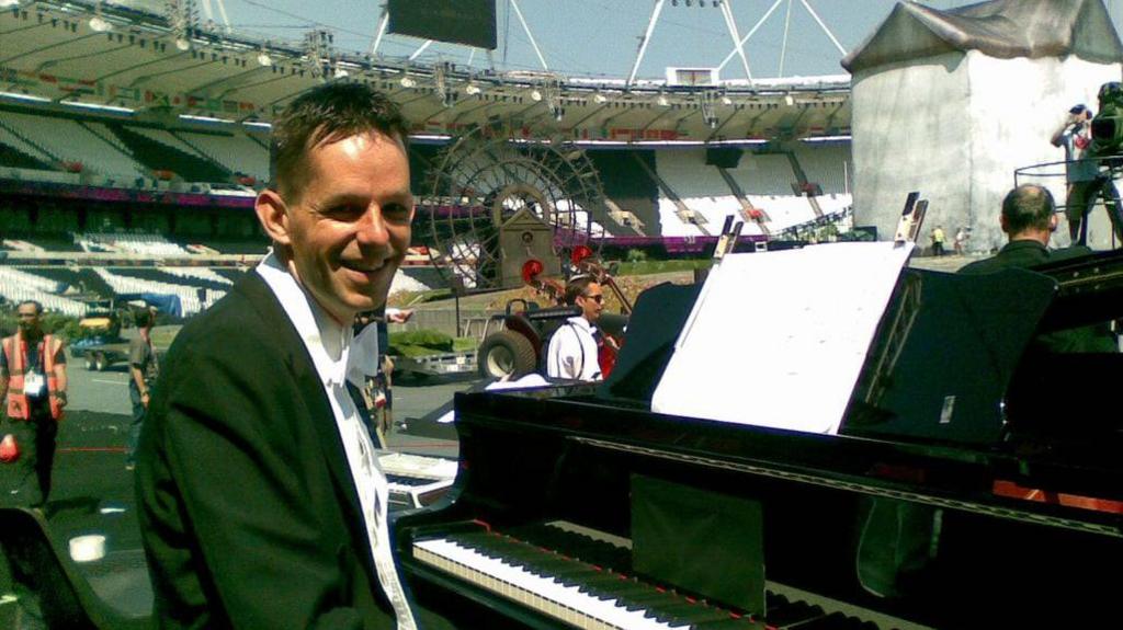 Iain Farrington in black tie sitting at a piano in a stadium. There are people and camera crews in the background as well as vehicles with trailers.