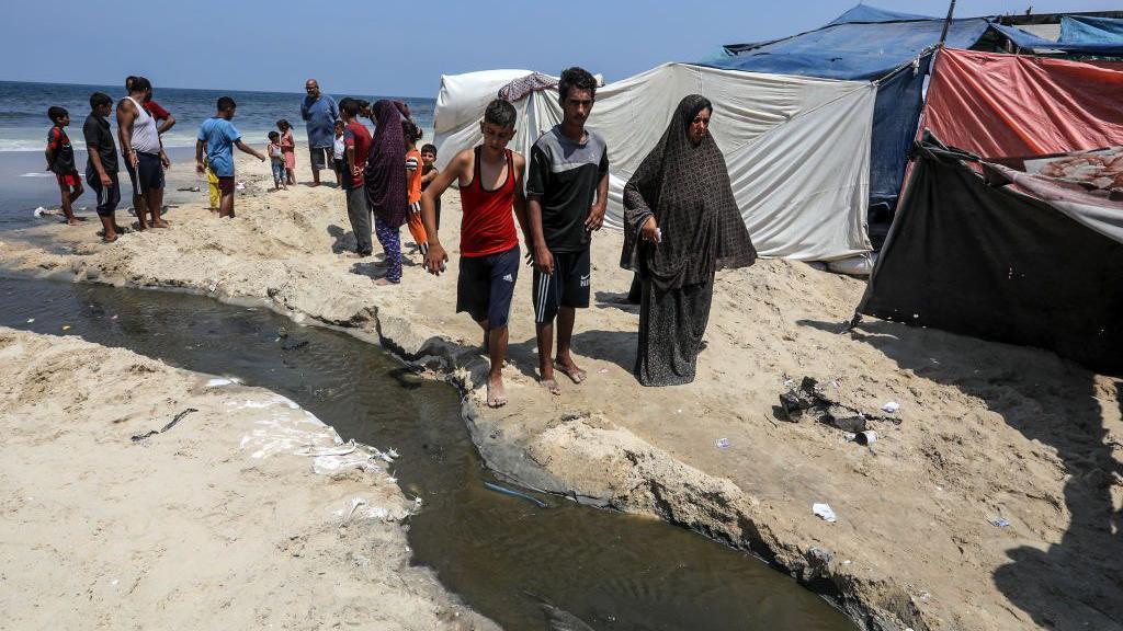 Displaced Palestinians stand next to a sewage-filled stream leading to the Mediterranean Sea on a beach near Deir al-Balah, central Gaza (19 August 2024)