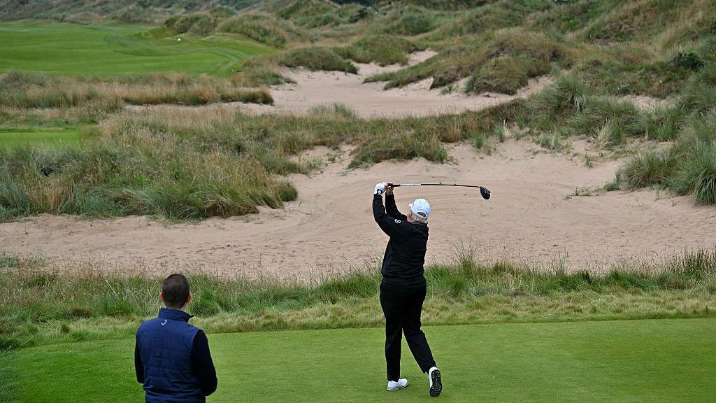 The back of Donald Trump wearing a white cap, black jumper and white golf glove mid swing with a golf club in the air.