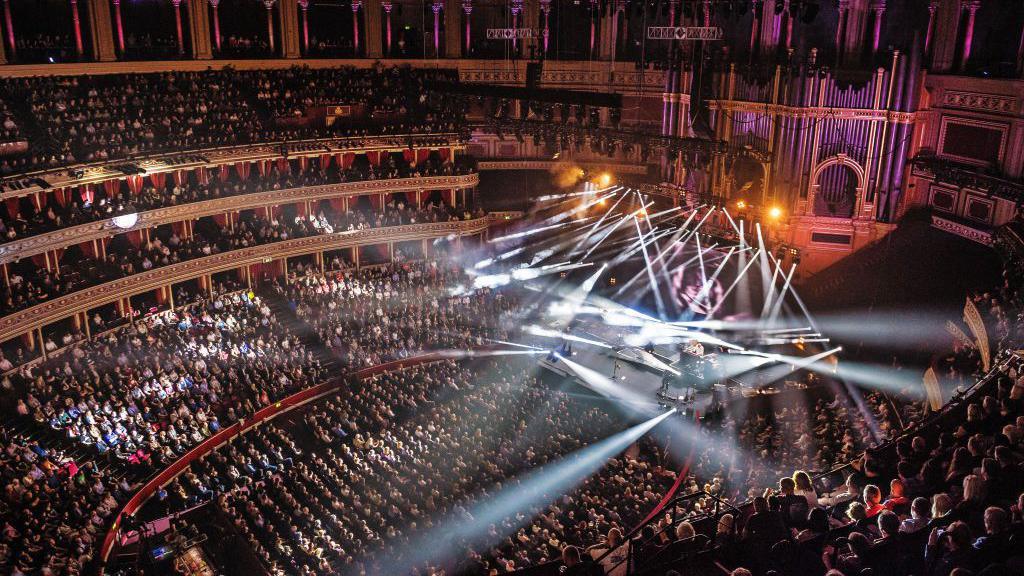 View of a packed audience inside the Royal Albert Hall during a live concert, with dramatic stage lighting and beams shining out into the crowd.